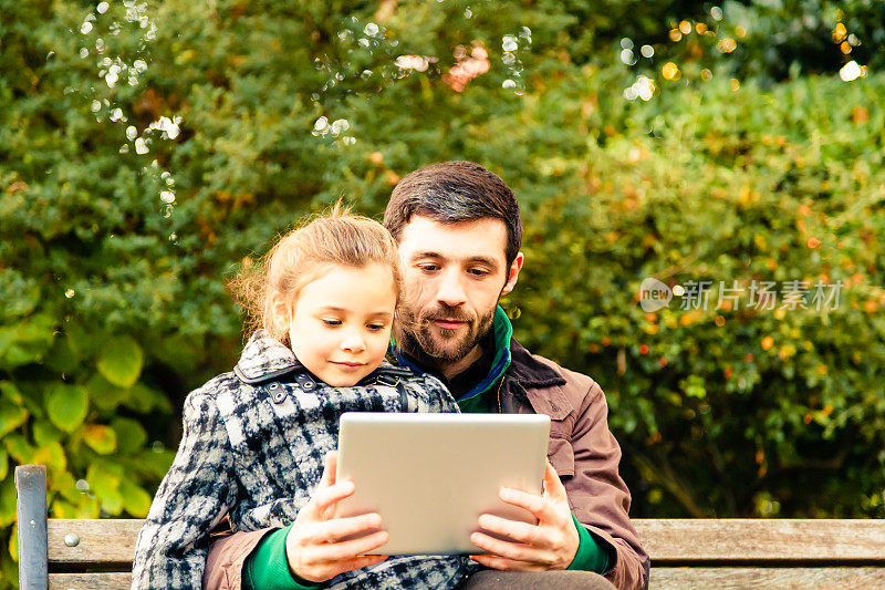 父亲和女儿坐在长凳上看平板电脑。