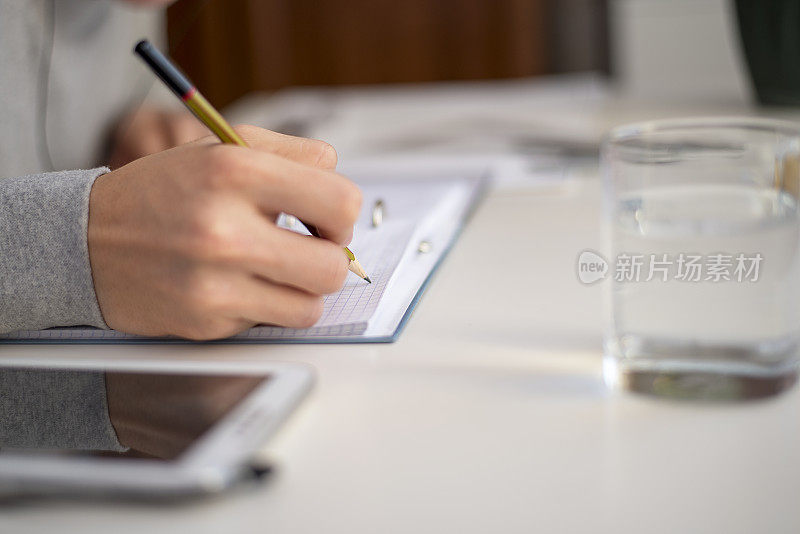 Unknown坐在一张白色的桌子旁，拿着一杯水和写字板，用铅笔在记事本上写字