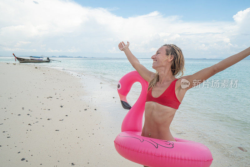 在泰国的岛屿上，一个年轻的女人在田园诗般的海滩上与充气火烈鸟展开双臂。人们旅游目的地有趣和酷的态度概念