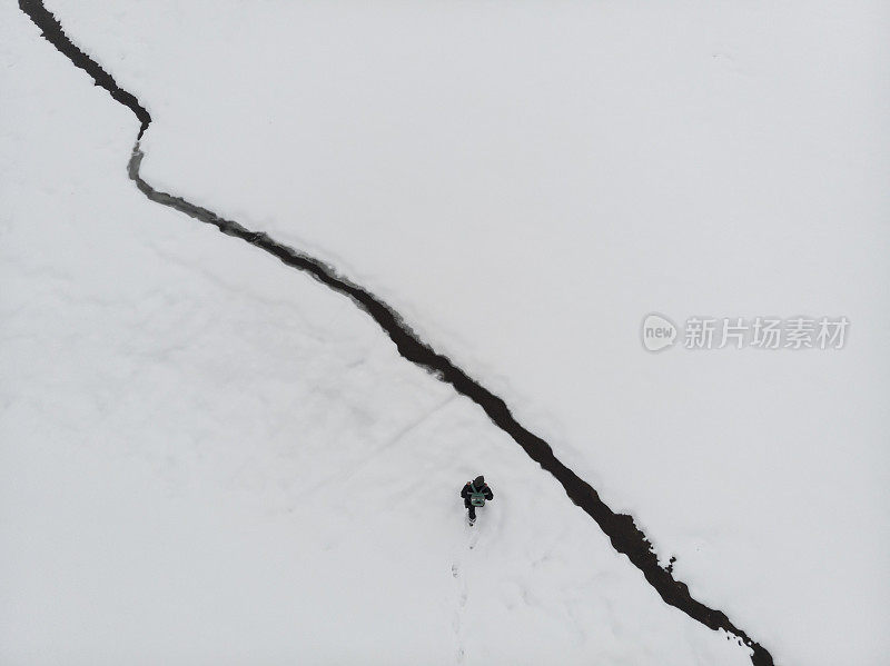 成年男子徒步行走在白雪覆盖的田野上，旁边是小水流。鸟瞰图，正上方。