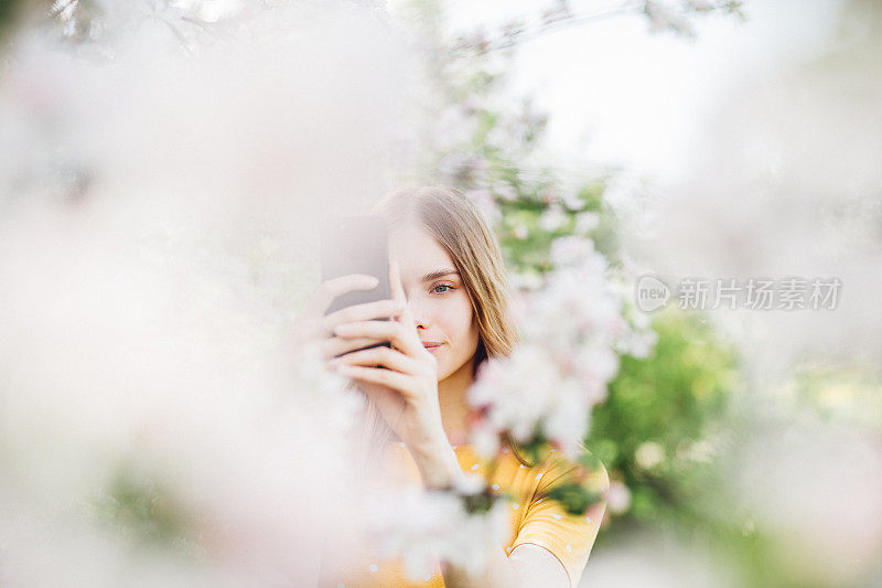 微笑的女人在盛开的树木，用她的手机。