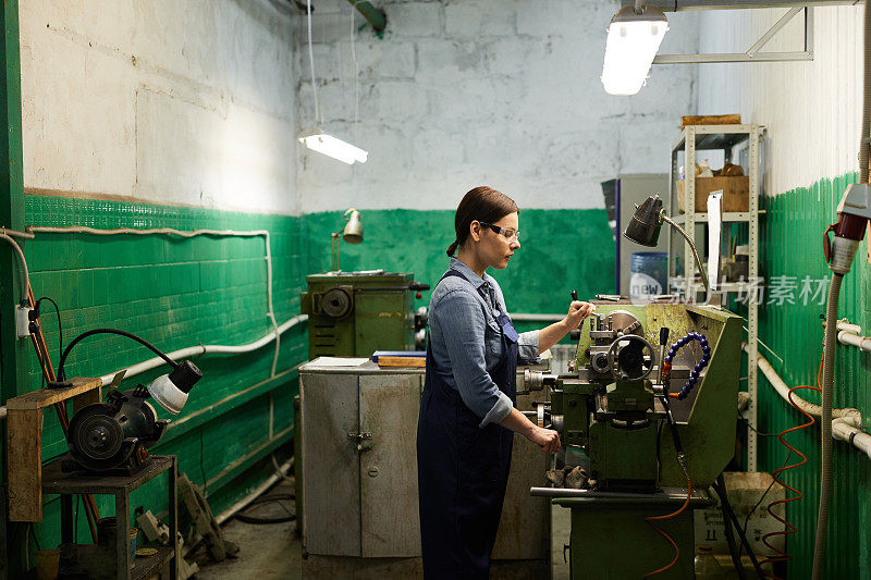 忙碌有经验的中年妇女戴着防护眼镜在工厂的手动车床上冲压金属