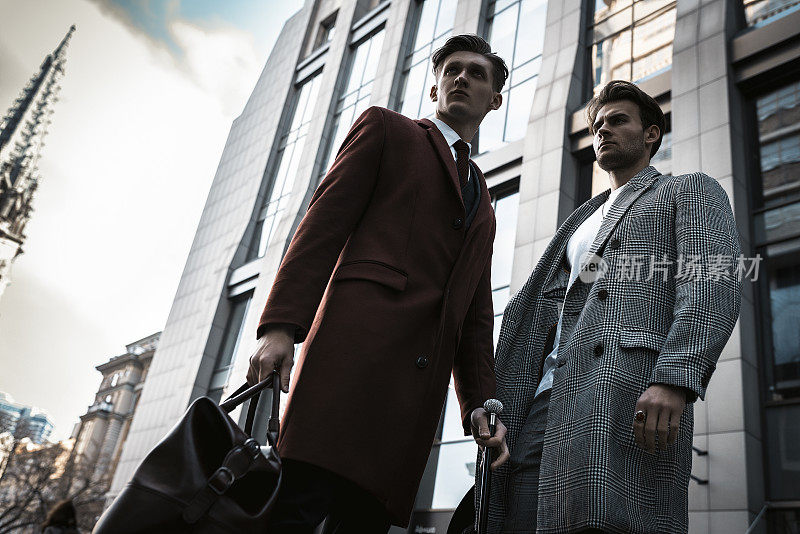 两个时尚男模特在城市街道上摆姿势。穿着在经典和休闲关闭。西装，外套，衬衫，毛衣，靴子和皮包。商业中心附近的男模测试
