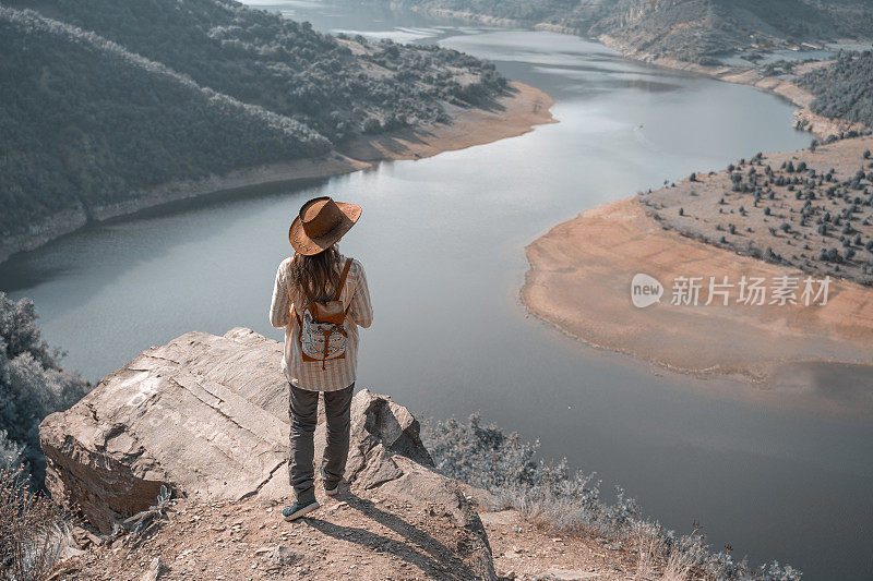 回归自然。独自旅行者在一个美丽的全景从高处俯瞰一条宽阔的弯曲的河流。一个快乐的年轻女游客漫步在大自然中。阳光明媚的户外享受。