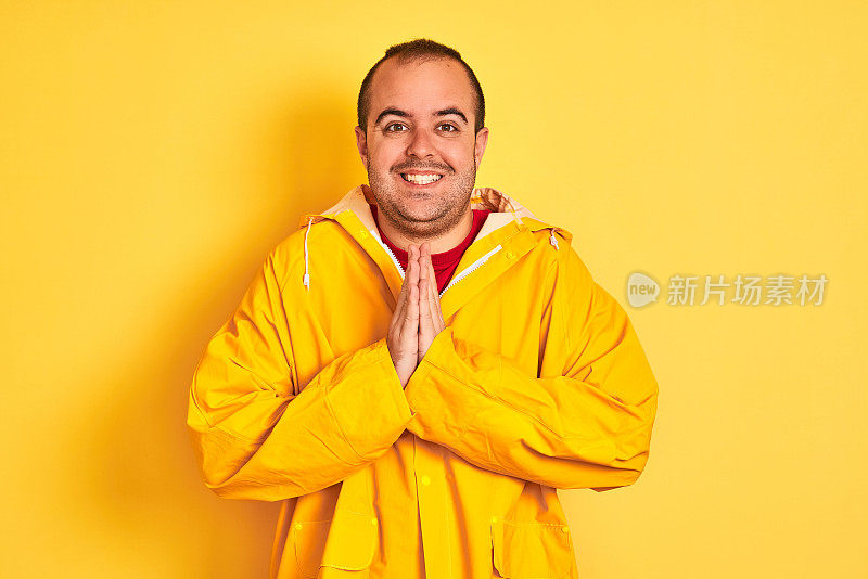 一个穿着雨衣的年轻人站在孤立的黄色背景上，双手祈祷，自信地微笑着请求宽恕。