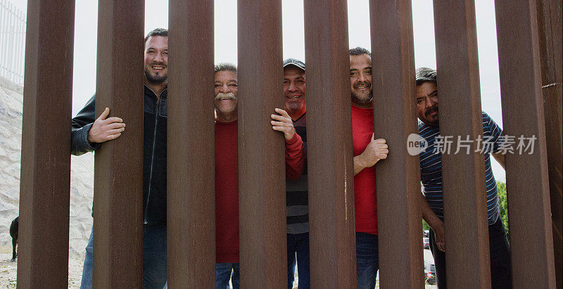 5名不同年龄的西班牙裔男子(在墨西哥一侧)穿过墨西哥和美国之间的钢板边境墙微笑