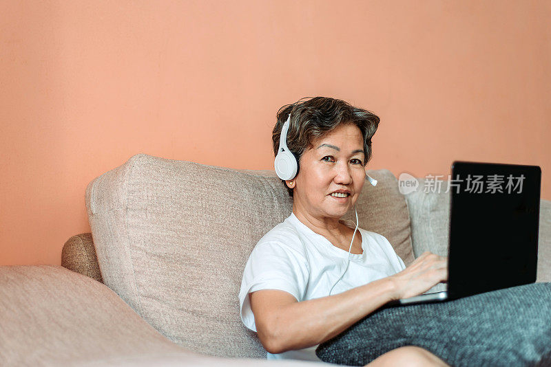 一名亚洲华裔高级女性坐在沙发上，戴着耳机使用笔记本电脑