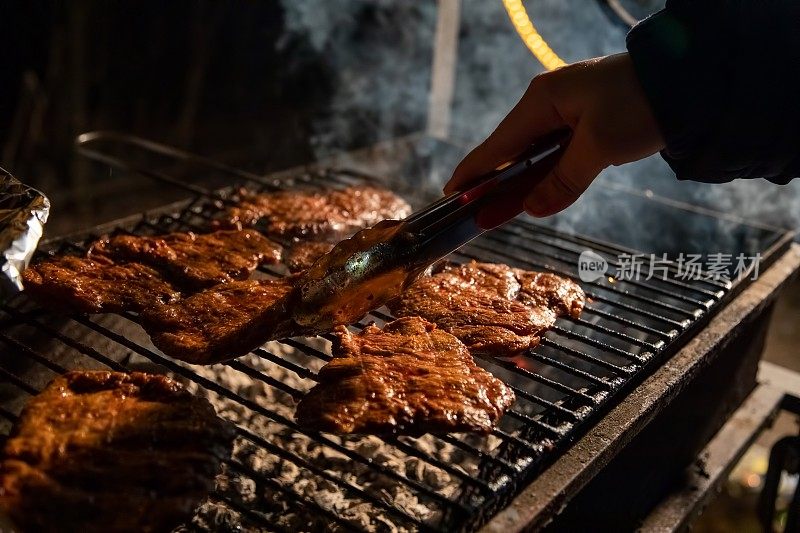晚上，一个烧烤架，美味多汁的牛排在篝火上烤着，一只手拿着钳子，把肉翻过来。特写,柔软的焦点。烟是强调