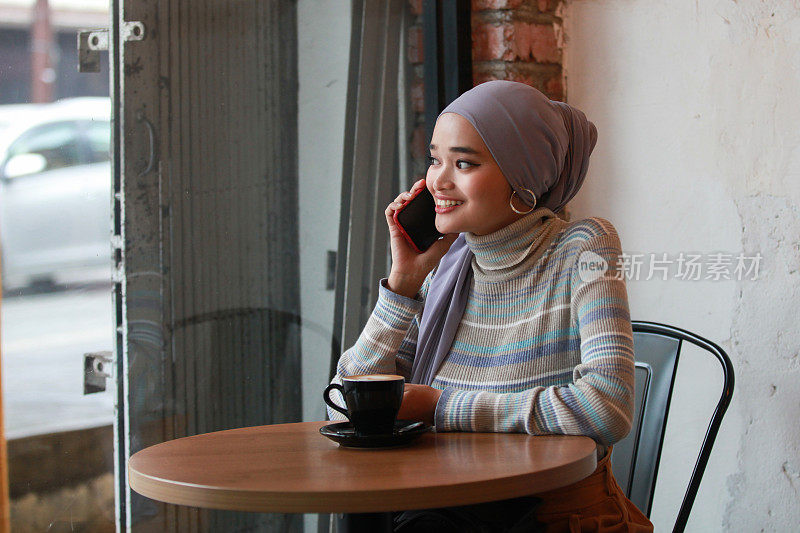 一个年轻的马来穆斯林妇女用手机和喝咖啡