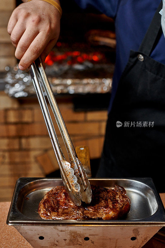 一位厨师在餐馆的厨房里用钳子把牛排翻过来。手和肉靠近