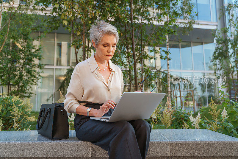 一位穿着黑色裤子的富有思想的女商人坐在商务中心附近的石凳上，在大腿上用她的笔记本电脑工作