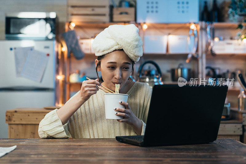 正面肖像，开朗的亚洲女孩，在舒适的家庭室内，在餐桌上吃方便面，同时在笔记本电脑上观看有趣的在线视频