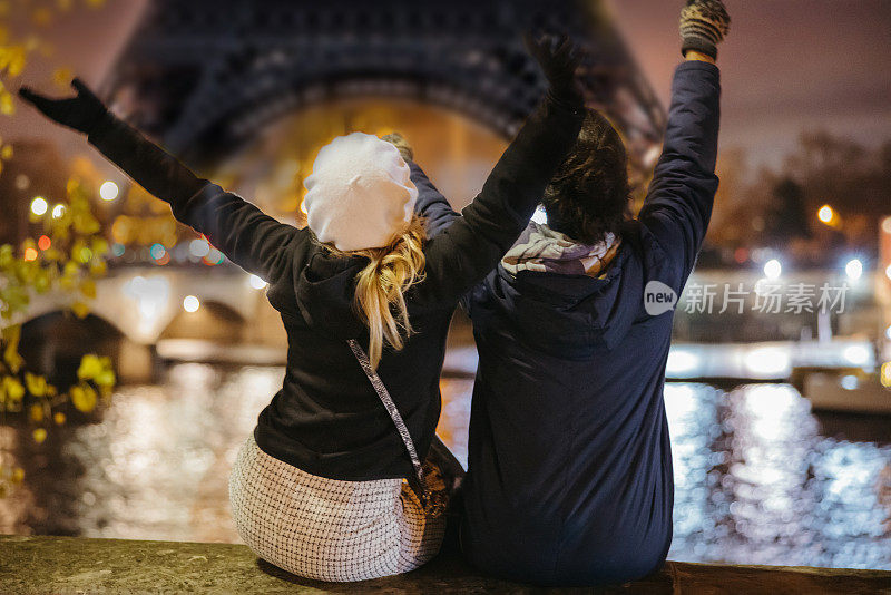 两个朋友坐在巴黎埃菲尔铁塔前塞纳河的城墙上，张开双臂