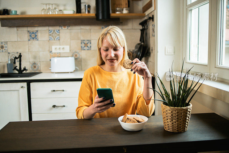 漂亮的金发女孩在家里吃饼干和使用智能手机