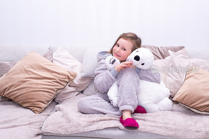一个七岁的白人女孩穿着家居服坐在柔软的沙发上，抱着一只大泰迪熊。