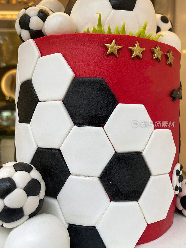 足球主题分层生日蛋糕的特写图像装饰在蛋糕板与黑色和白色六角形翻糖糖衣镶嵌瓷砖与红色背景，足球和绿叶草翻糖糖衣的叶片，重点在前景