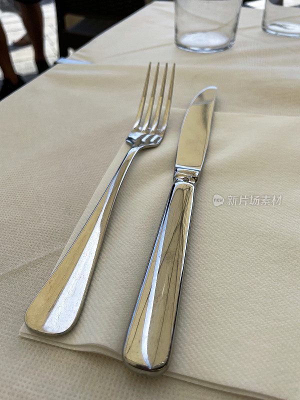 全画幅的露天餐厅餐桌布景，米色桌布上的不锈钢刀叉餐具，空的水杯，抬高的视野，聚焦于前景