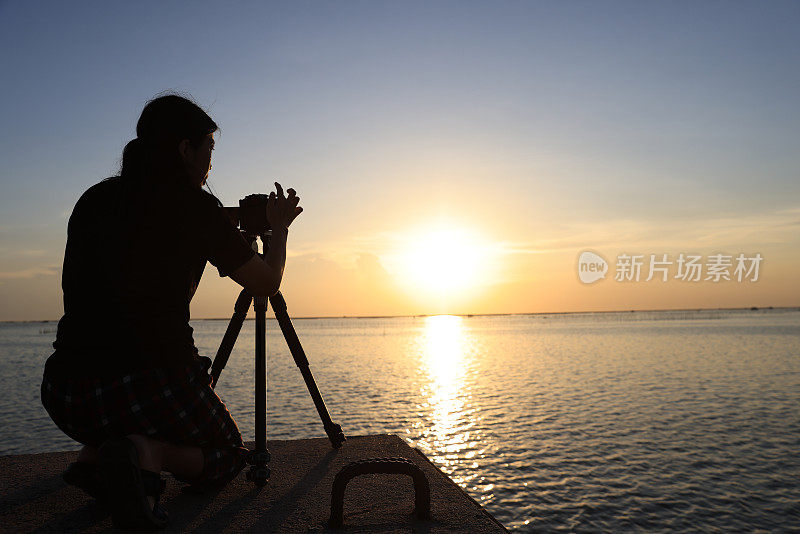 一个女人在夕阳下拍照的剪影。摄影师用剪影在桥上拍摄日落照片。准备好相机和三脚架，准备在桥上拍一张日落照片。