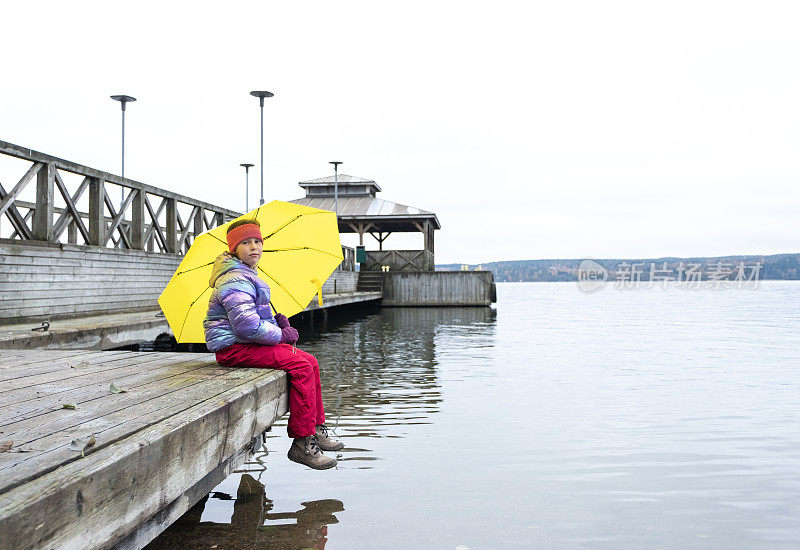一个8岁的白人女孩坐在木码头上，拿着一把黄色的雨伞。秋天走在湖岸。