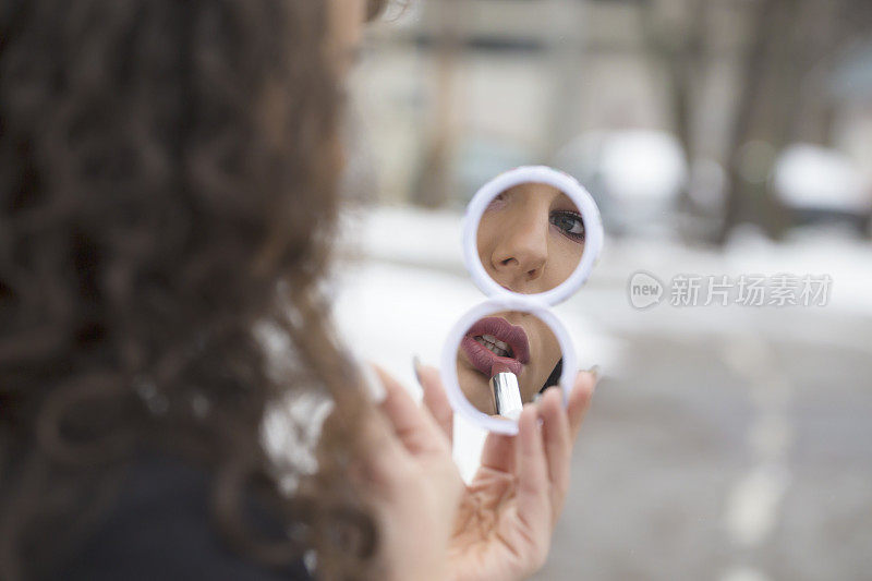 美丽的蓝眼睛卷发女孩在冬天的室外看着镜子涂口红的肖像