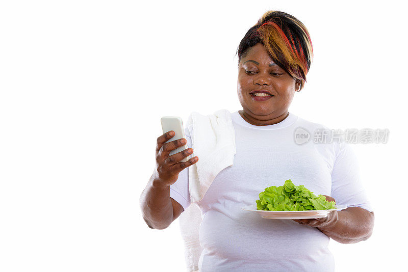 摄影工作室拍摄到一名快乐的非洲大码黑人妇女微笑着使用手机，手里拿着盛在白色盘子里的生菜，并在视频中聊天