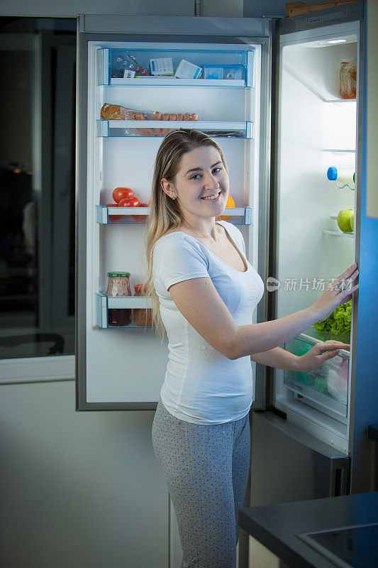 微笑的女人在晚上在开放的冰箱摆姿势的肖像
