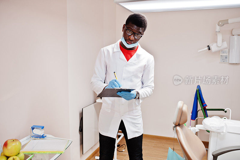 戴眼镜和面具的非裔美国男医生在诊所里摆姿势，手里拿着黑色的写字板，写着什么。