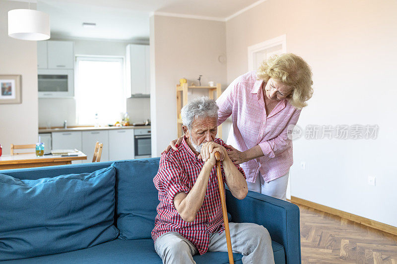 悲伤的退休丈夫拄着拐杖坐在客厅里，旁边是老妻子。老夫妇看着对方，手牵着手在家里