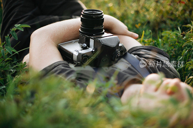 时髦的潮人与电影相机躺在绿色的草地上，放松在阳光灿烂的草地在大气的日落。帅哥在大自然中休息，在山中享受夜晚