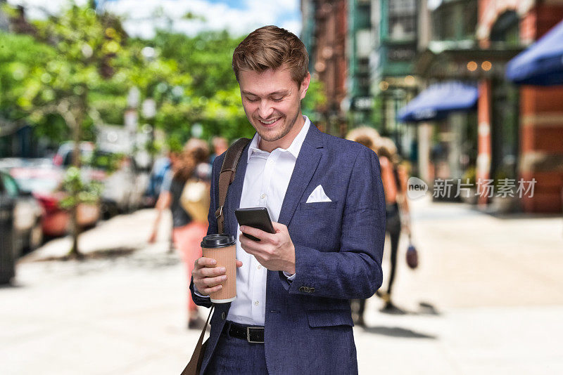 站在离焦背景的年轻男性商人，背着肩包，拿着咖啡杯，在户外使用智能手机