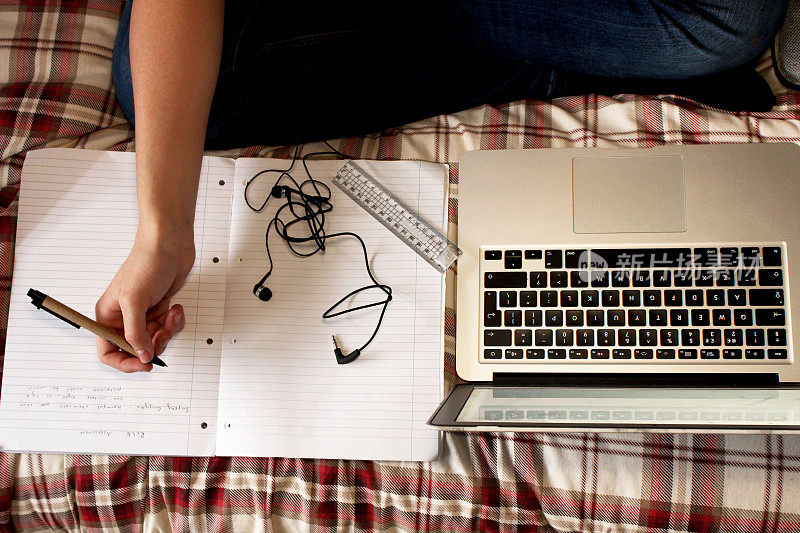 照片中的学生盘腿坐在床上，用笔记本电脑工作，用圆珠笔、尺子、耳机、高架视野在横线笔记本上写字
