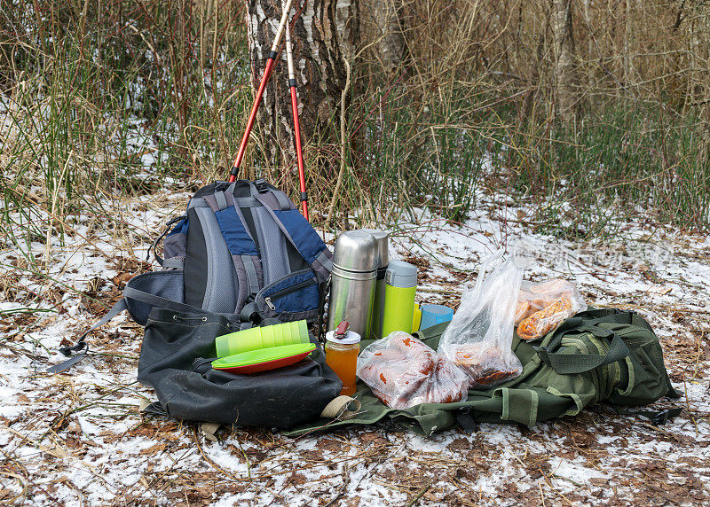 雪地背景和野餐用品，背包，杯子，保温瓶和各种菜肴在地面上，游客午餐在大自然的冬天