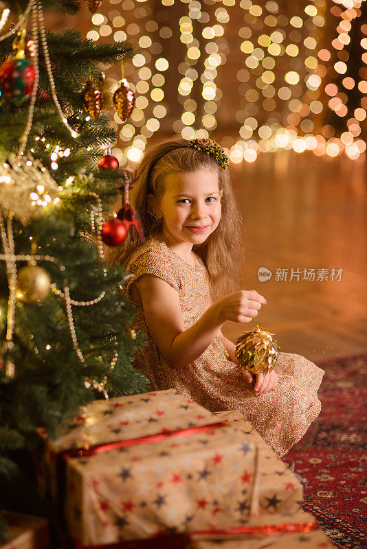 女孩穿着漂亮的衣服，坐在一棵有装饰的圣诞树下，靠近礼品盒。手里拿着一个玩具
