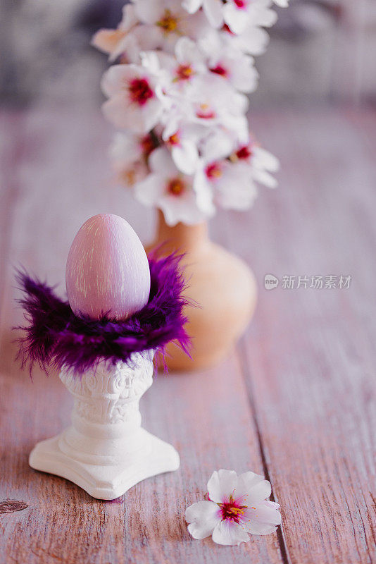 简单而美丽的静物，一个紫色的复活节彩蛋，上面有紫色的羽毛，上面有一个白色的小柱子，还有一个插着杏花树枝的花瓶