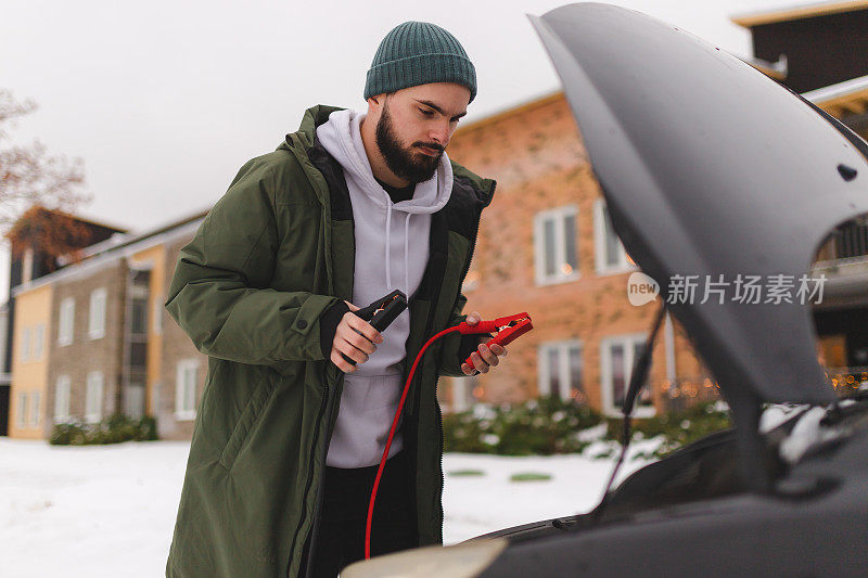 穿着冬装的年轻人在冬天用电池电缆启动他的汽车