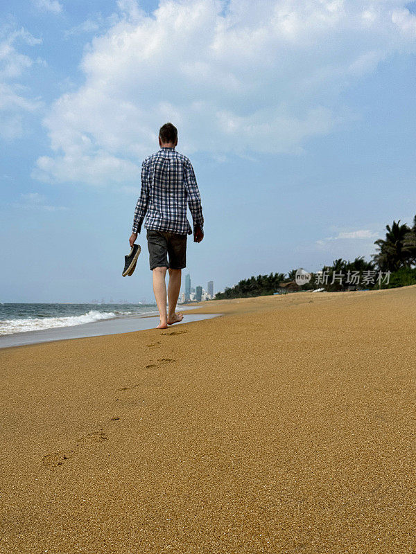特写图像，无法辨认的男子沿着热带拉维尼亚山海滩走到远处，在沙滩上留下脚印，在斯里兰卡科伦坡度假，穿着短裤和方格衬衫，拿着沙滩鞋，后视图
