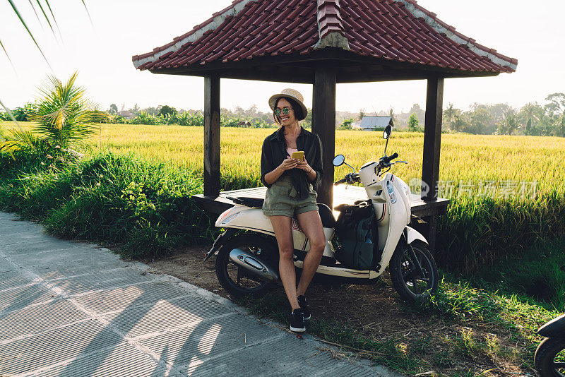 长着牙齿的女旅行者，手里拿着手机，在独自旅行时骑着复古摩托车，快乐的高加索博主在印度尼西亚使用手机小工具和老式交通工具