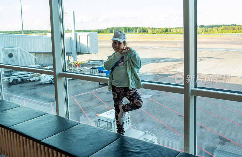 一个孩子，一个白人女孩，在机场一个巨大的窗户的背景下，看着镜头，展示了一个爱心的手势。
