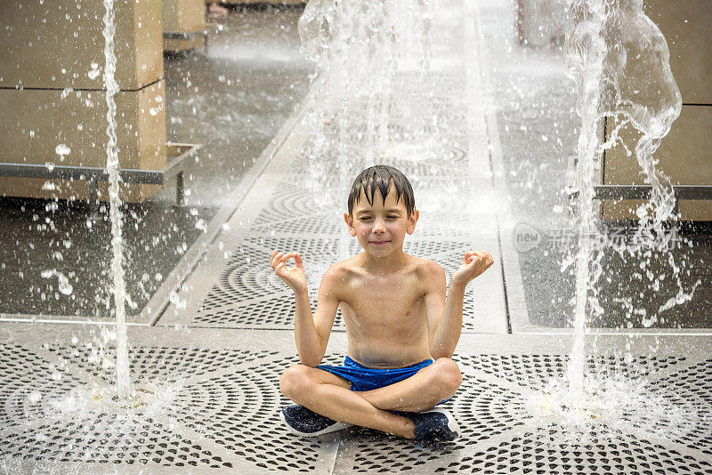 男孩在喷泉里冥想，发现禅。在炎热的夏日，一个孩子在城市喷泉边玩耍。快乐的孩子们在喷泉里玩耍。夏天的天气。积极休闲，生活方式和度假
