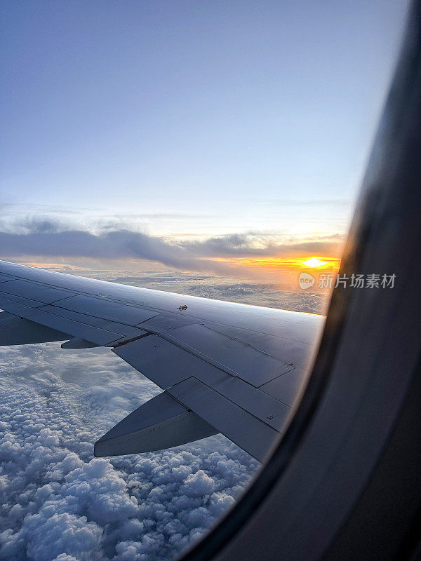 从飞机窗口看到的图像，飞机机翼在云层之上，蓬松的白云在海上，机翼板条，橙色的日落，重点在前景