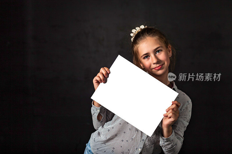 童女宣传白色海报模型空白框，在黑色背景下摆姿势，微笑着看着镜头。孩子拿着空模板，手里拿着空白的横幅。广告的概念。复制广告文本空间
