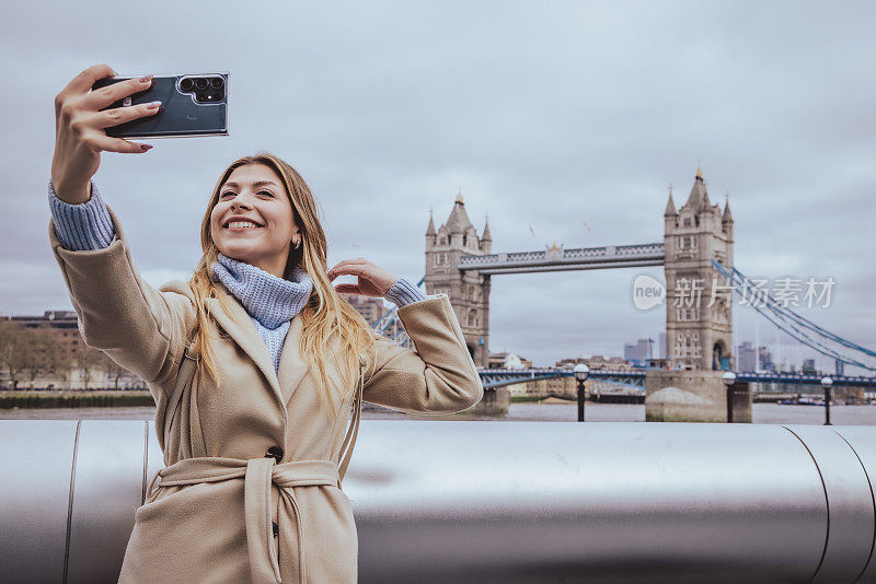 在英国伦敦塔桥前，一位30多岁的金发美女微笑着用智能手机自拍。这张照片很有创意