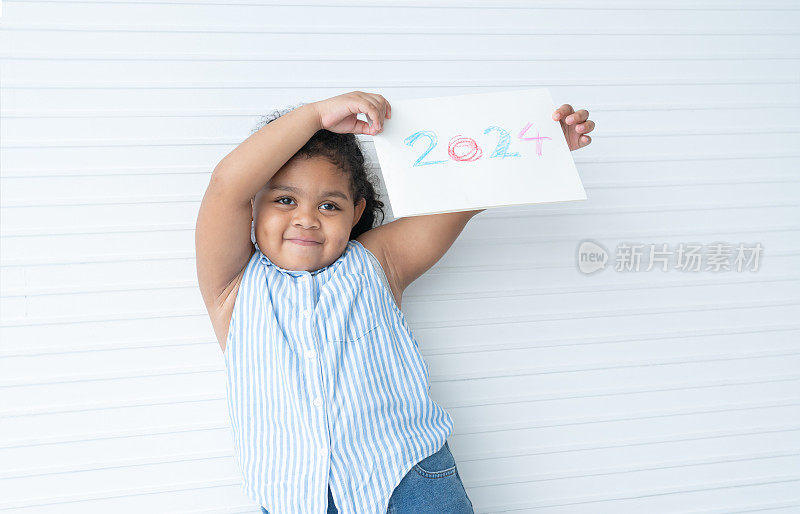 小可爱的非洲小女孩拿着手工制作的卡片，上面写着“2024”，微笑着，在白底上展示着一张欢快的脸。孩子们庆祝新年的表情