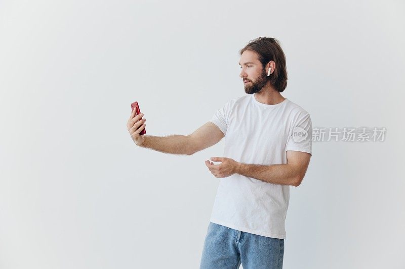 一个留着胡子的博主，穿着白色t恤，拿着手机和无线耳机，在白色背景下进行在线视频通话