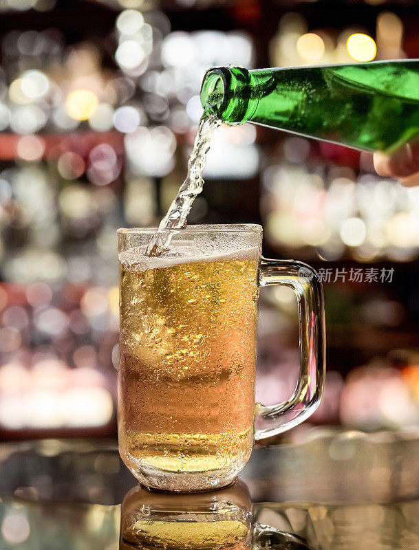 啤酒从绿色啤酒瓶倒进玻璃大酒杯的特写图片，啤酒杯上覆盖着凝结的气泡，在啤酒上形成泡沫的泡沫头，木制吧台，酒吧背景，焦点在前景上