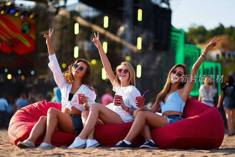 日落时分，女士们坐在豆袋椅上敬酒，海滩音乐节正如火如荼地进行着。三个朋友一起享受欢乐时光，无忧无虑的夏日氛围。充满笑声、欢呼声和海边氛围的户外活动。