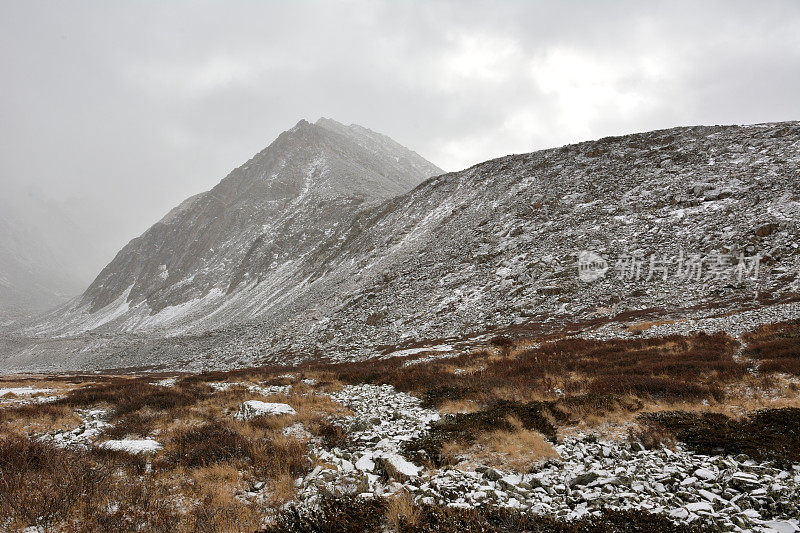 高山陡峭的斜坡上有松散的石头，向下延伸到一个风景如画的山谷，洒着第一场雪。
