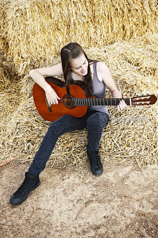 美丽的乡村女孩坐在稻草上弹吉他，微笑着