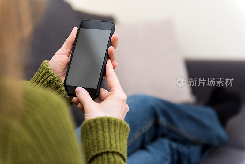 一个金发的年轻女子正坐在家里，手里拿着手机，看着一个空白的智能手机。