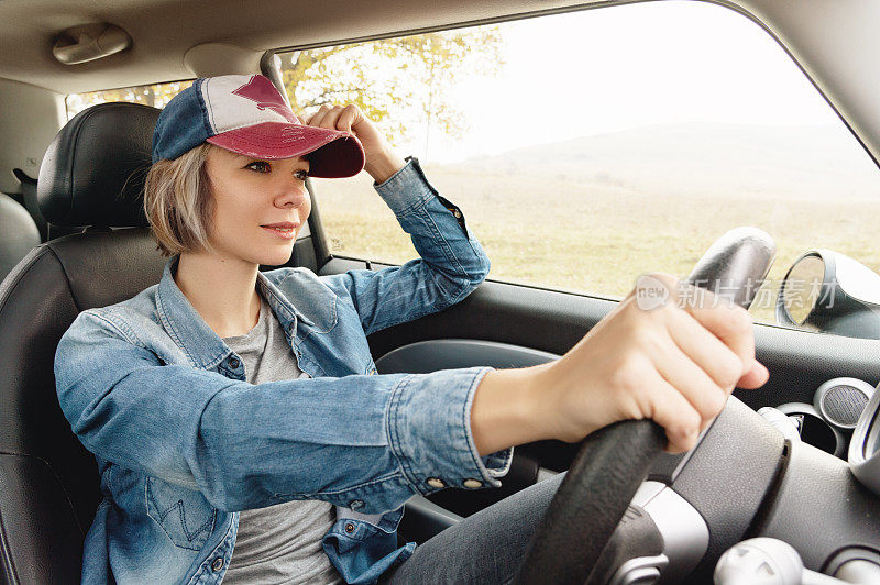 快乐漂亮的女孩在牛仔裤衣服和帽子微笑在她的汽车方向盘